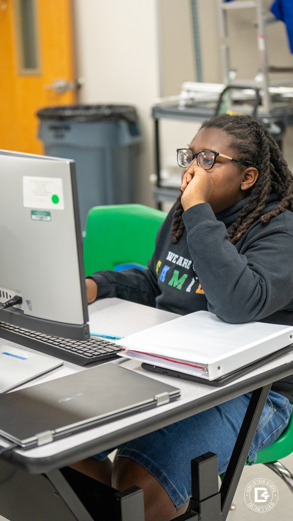 A student wearing glasses and a black hoodie labeled “We Are Family” focuses intently on a computer screen, resting their hand on their face. A large binder and laptop are on the desk.