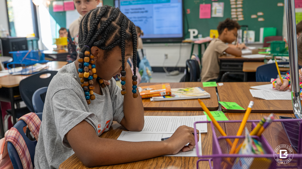 A student with colorful beads in her braids studies her worksheet at her desk, with other students working in the background.