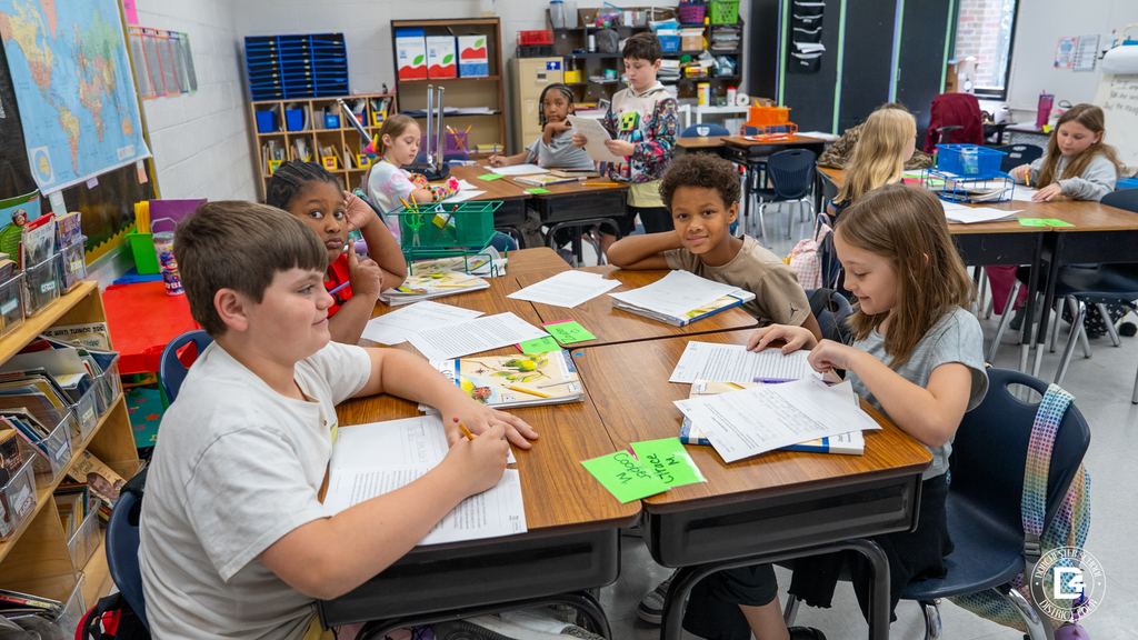 A group of elementary students sit together at a table, reading and writing on worksheets. Some look up toward the camera while others stay focused on their papers.