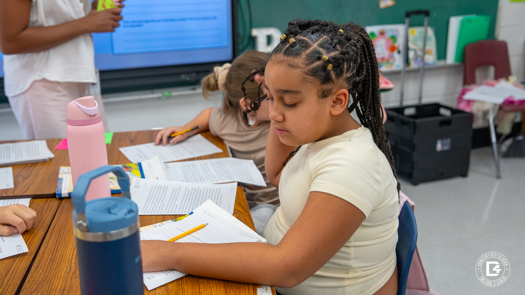 A student with braided hair and beads concentrates on her worksheet, pencil in hand, with a water bottle and papers spread across the desk.