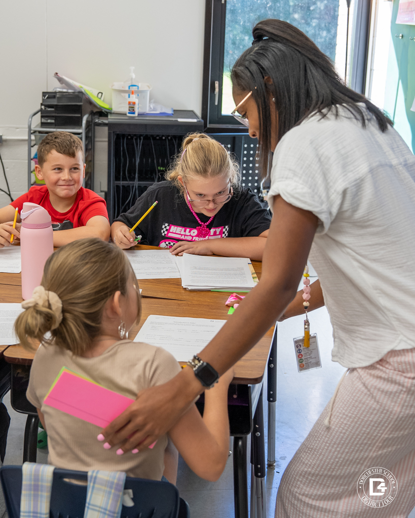 A teacher leans down to assist students at a table. Two children smile while another focuses on writing, and a student in the foreground listens with a pink water bottle nearby.