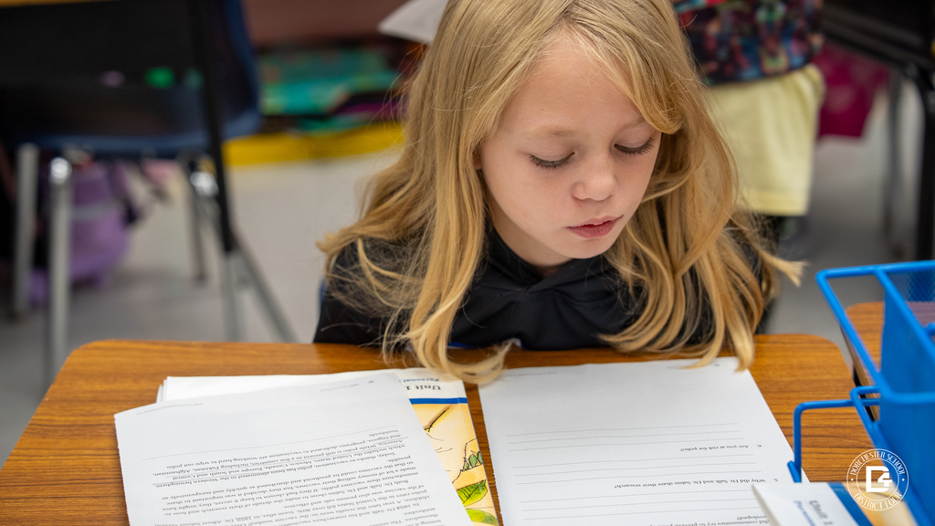 A young girl with long blonde hair reads intently at her desk, her papers and book laid out in front of her.