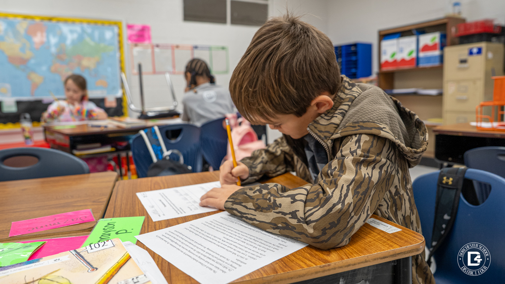 A student in a camouflage hoodie writes carefully on his worksheet, leaning over the desk with focus.