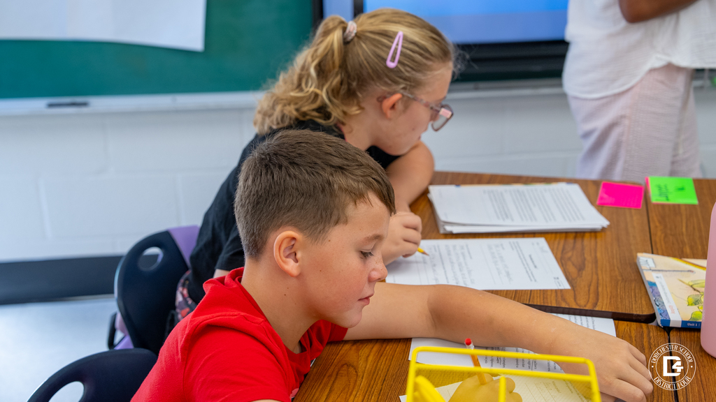 Two students lean over their papers at a shared table, writing with pencils while listening to their teacher at the front of the class.