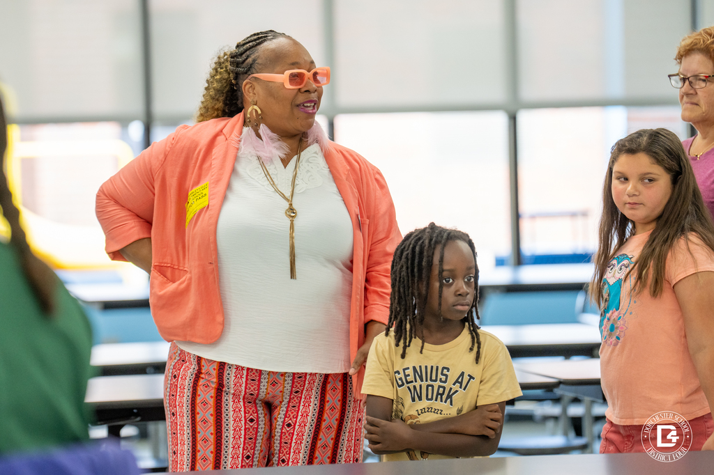 A woman in a coral jacket with bright orange glasses stands in a cafeteria with children nearby. One child wears a shirt that says “Genius at Work.”