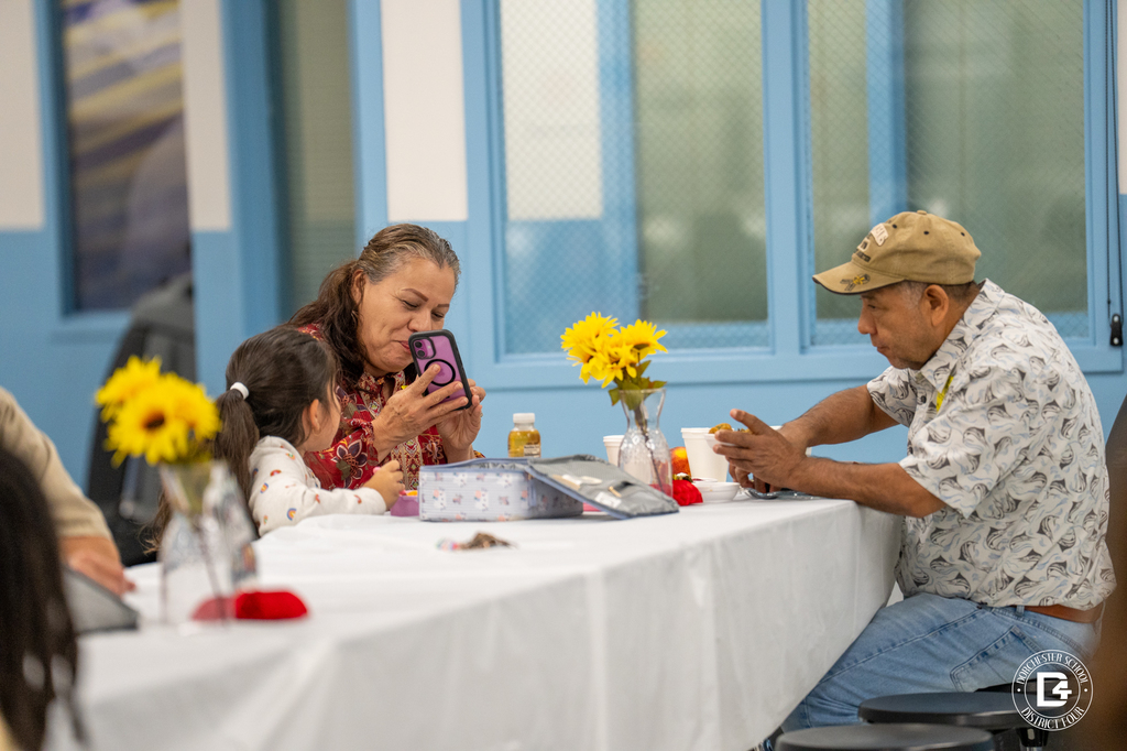 A woman sits at a decorated table with a young child, smiling at her phone, while a man in a tan cap sits across from them eating. A vase of bright yellow flowers sits in the middle of the table.