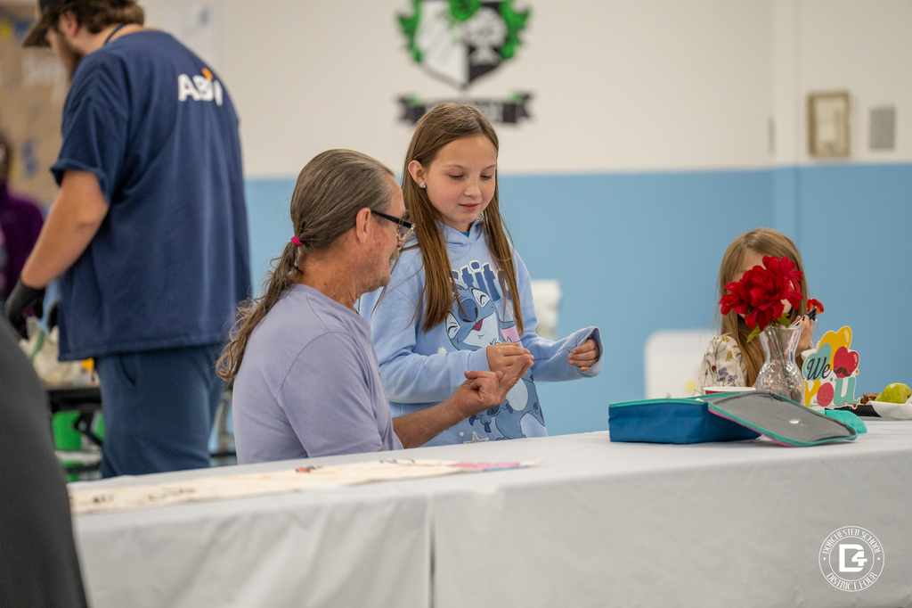 A man with long hair tied in a ponytail sits at a table talking with a young girl in a light blue hoodie. A vase of red flowers decorates the table.