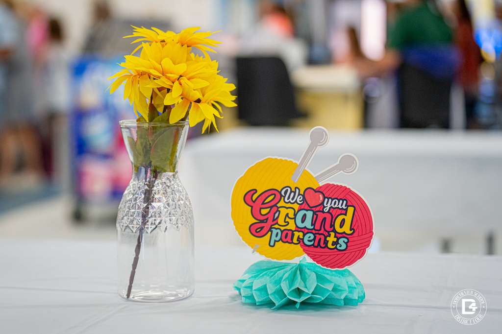 A clear vase filled with yellow flowers sits on a table next to a colorful sign that reads “We love you Grandparents.”
