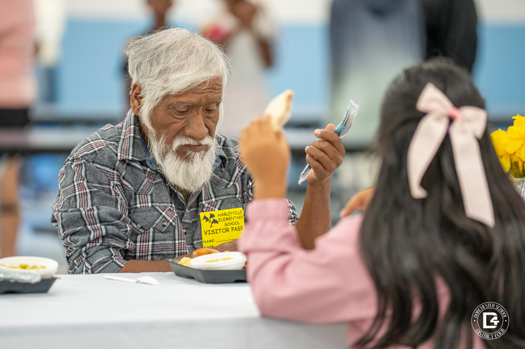 An older man with a white beard and plaid shirt eats at a cafeteria table while a child with a pink bow in her hair sits across from him. He is wearing a visitor pass from Harleyville Elementary School.