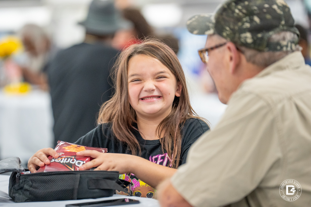 A young girl with long brown hair smiles widely at the camera while holding a bag of Doritos. She sits at a cafeteria table across from an older man wearing glasses and a camouflage hat.