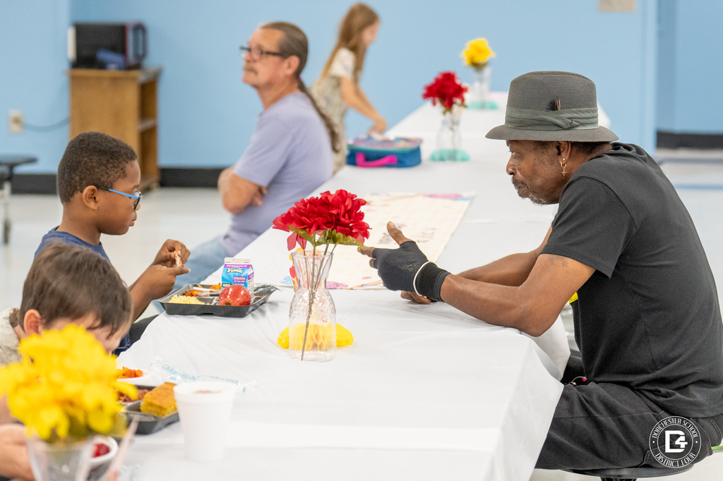 A man wearing a gray hat sits across from a young boy in glasses, sharing a meal at a long cafeteria table decorated with flowers. Other families can be seen in the background.