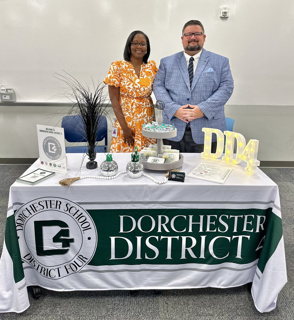 Two Dorchester School District Four representatives stand behind a decorated display table at the Career Fair. The table is covered with a white and green DD4 tablecloth featuring the district logo. On top are brochures, a sign, decorative items, candy, and light-up letters spelling “DD4.” The woman is wearing an orange floral dress and the man is dressed in a light blue suit with a striped tie.