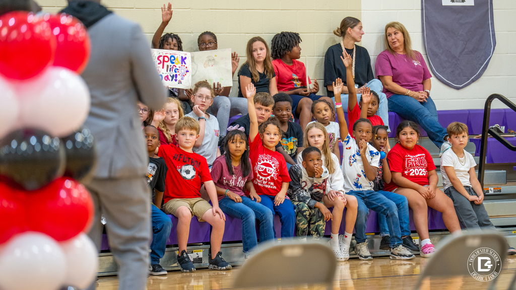 Michael White speaks at the front of the gym while a group of CHES students in the bleachers raise their hands. A handmade “Speak Life Day” poster can be seen in the background.
