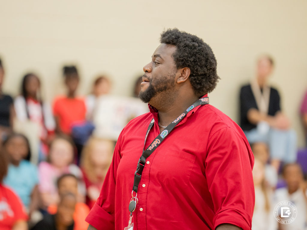 A CHES staff member in a red shirt stands in the gym, looking to the side, with students in the background.