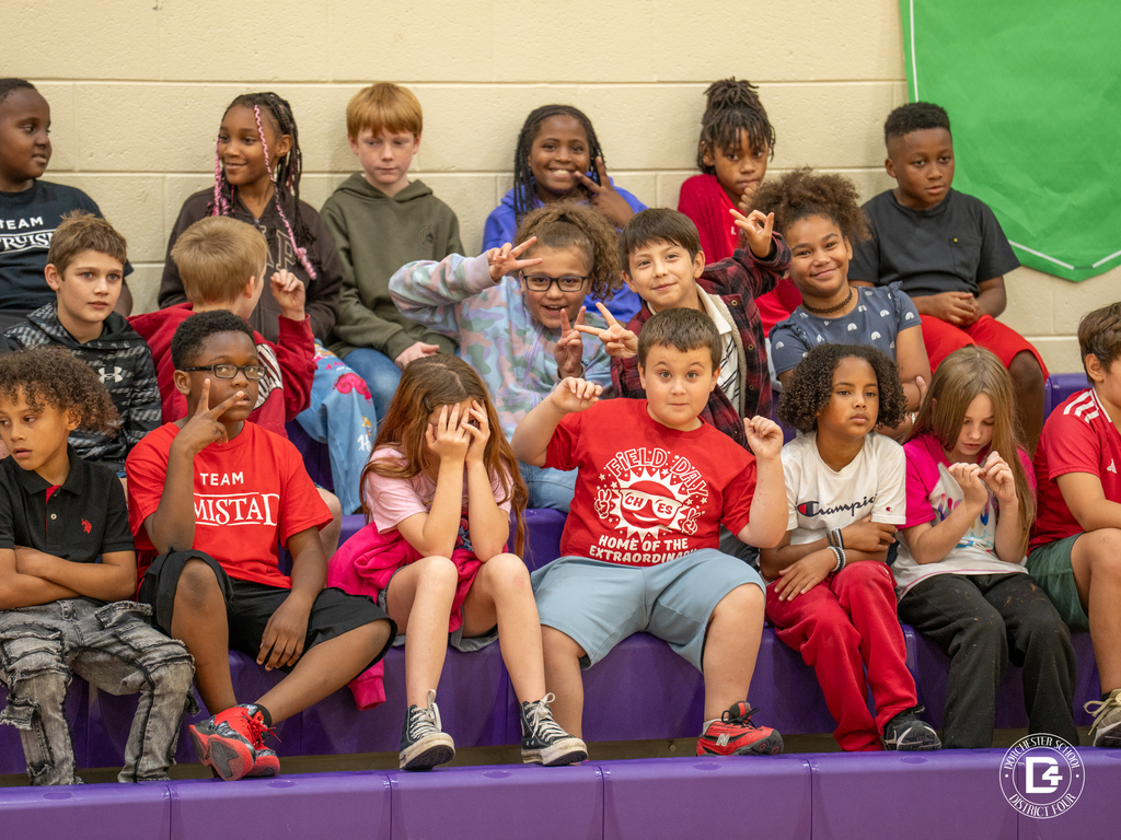 A group of upper elementary students sit in the bleachers making peace signs, smiling, and posing for the camera, while others look on during the program.