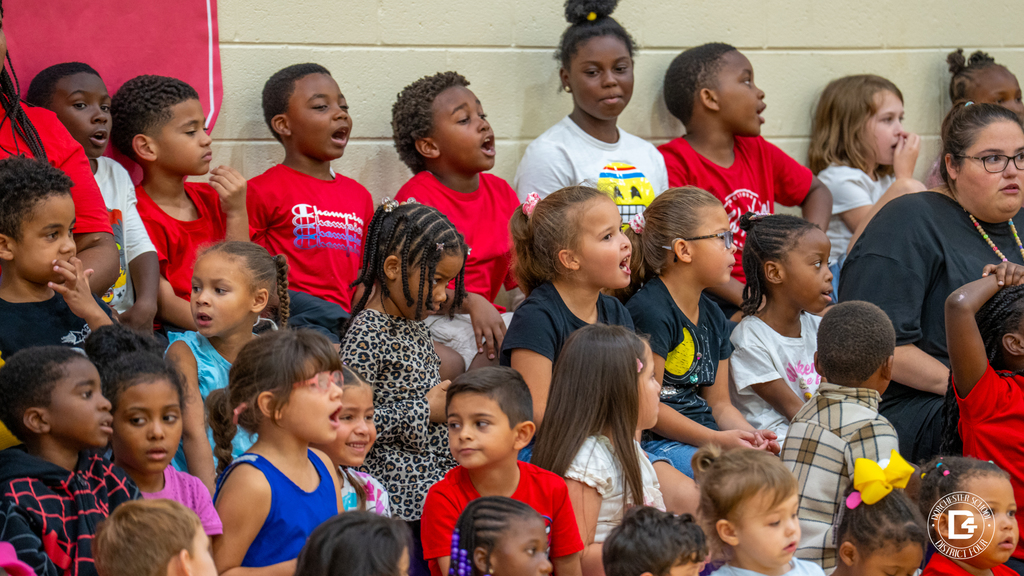 A section of the bleachers filled with elementary students wearing red, black, and white shirts. Many students are smiling and singing along during the event.