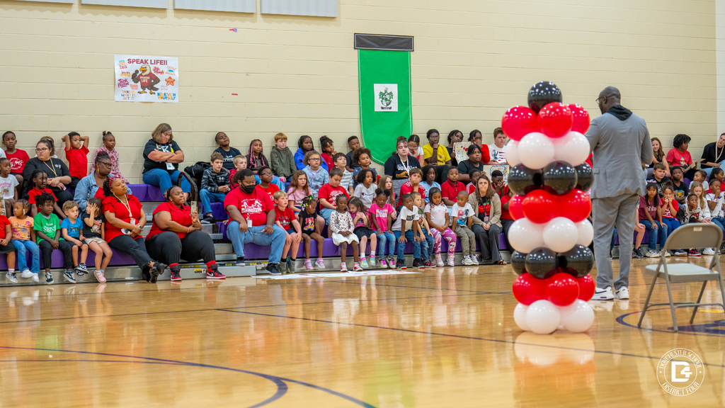 A wide-angle view of the event, showing Michael White addressing the student body. Rows of students and teachers fill the bleachers, with red, black, and white balloon columns on the gym floor.