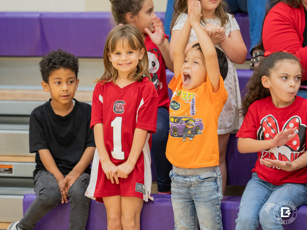 Three children in the bleachers. One child cheers with both arms raised, another smiles at the camera, and the third watches attentively.