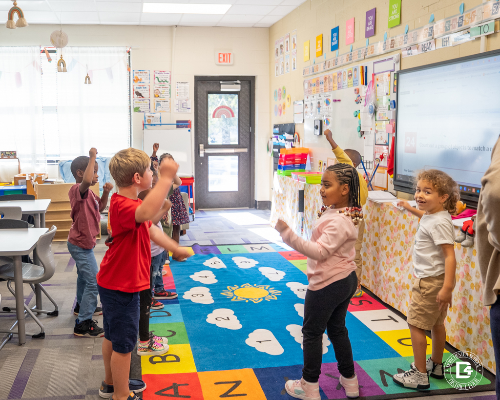 Kindergarten students stand on the classroom rug, moving their arms as they count together. One student smiles toward the camera while participating in the activity.
