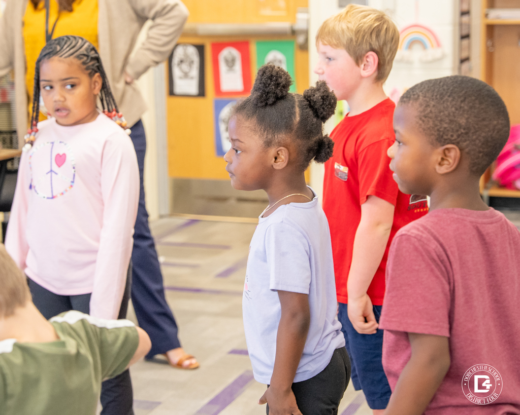 Four kindergarten students stand side by side, watching attentively as they participate in their classroom warm-up activity.