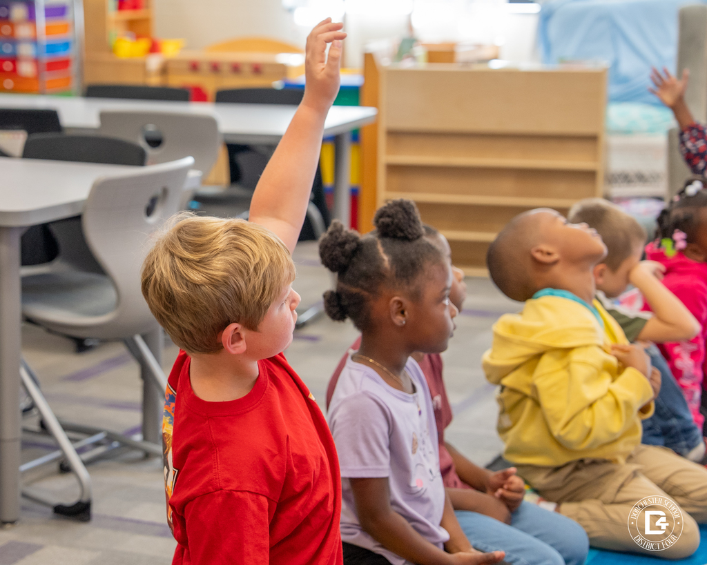 Several kindergarten students sit in a row on the classroom floor. One boy in a red shirt raises his hand while other students look up toward Mrs. Rudd.