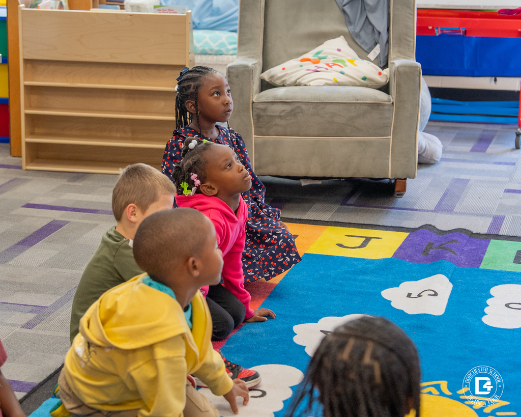 Kindergarten students sit attentively on a rug decorated with numbers and letters, watching Mrs. Rudd at the front of the classroom.