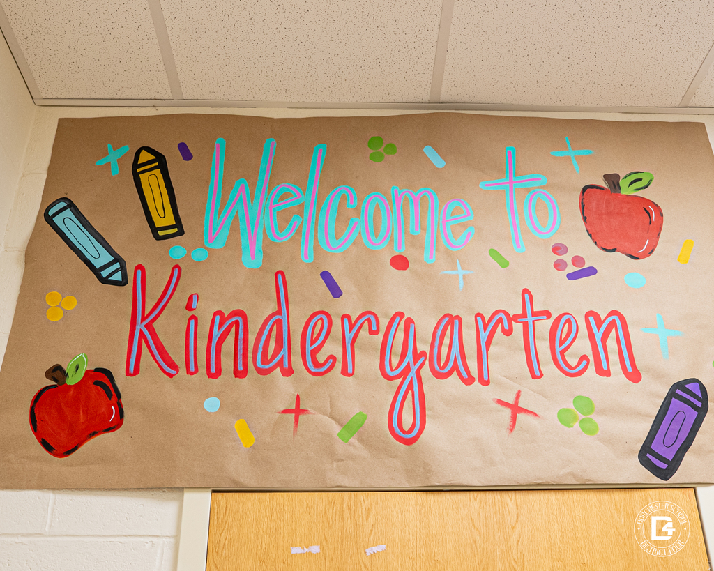 A colorful classroom banner reads “Welcome to Kindergarten” with bright drawings of crayons, apples, and confetti-like shapes on brown paper above a doorway.