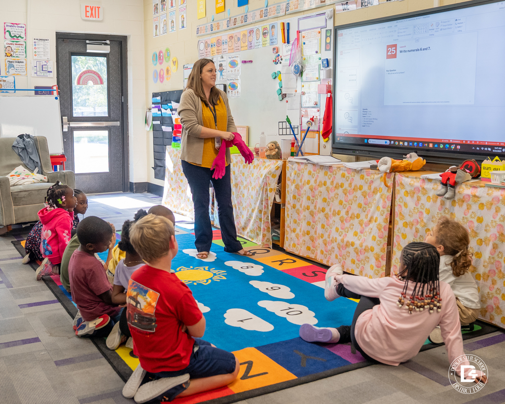 Mrs. Rudd stands at the front of the classroom holding pink gloves, speaking to her kindergarten students who are sitting on a colorful rug. A large screen behind her displays a math lesson about writing numerals.