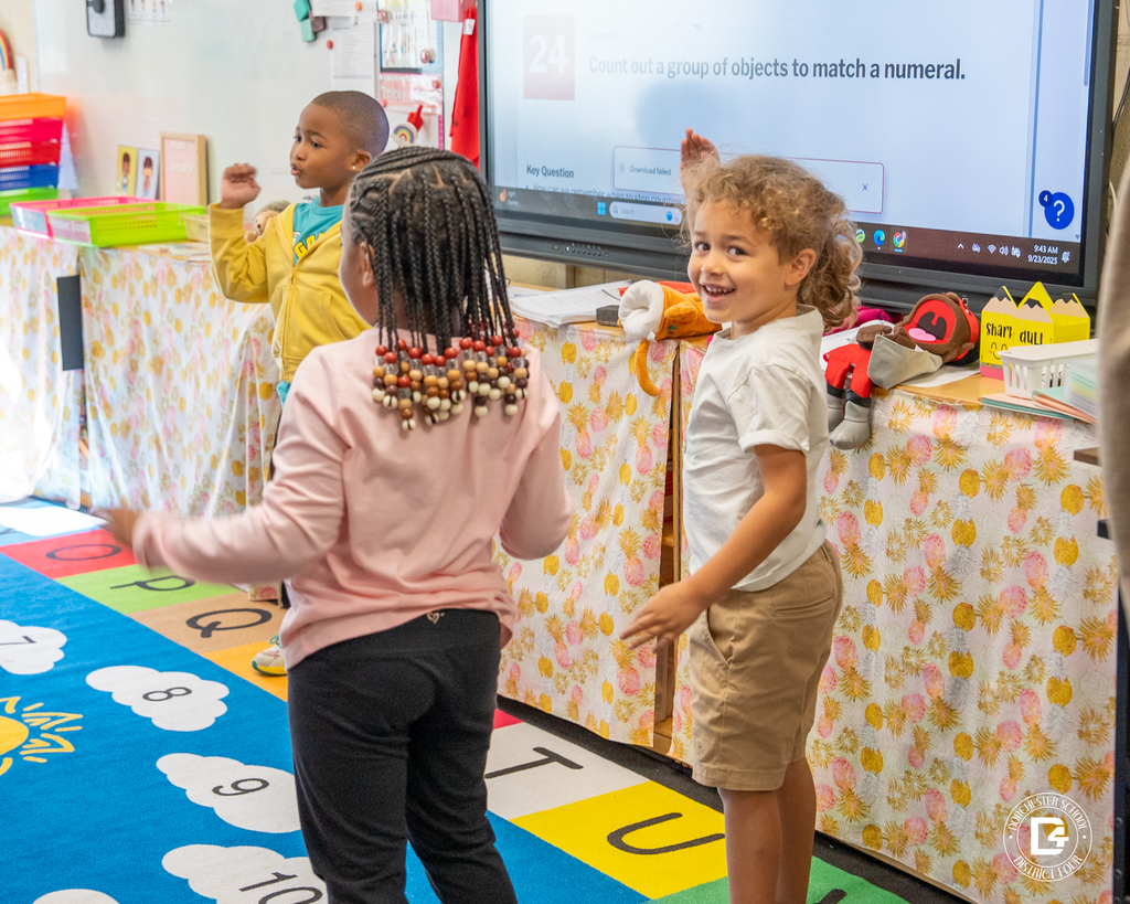 Two kindergarten students stand near the classroom board smiling and moving their arms during an interactive lesson.