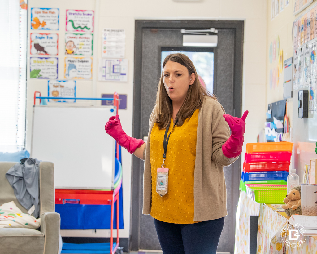 Mrs. Rudd, wearing a yellow shirt and beige sweater, gestures with both hands while wearing bright pink gloves as she teaches her kindergarten students.