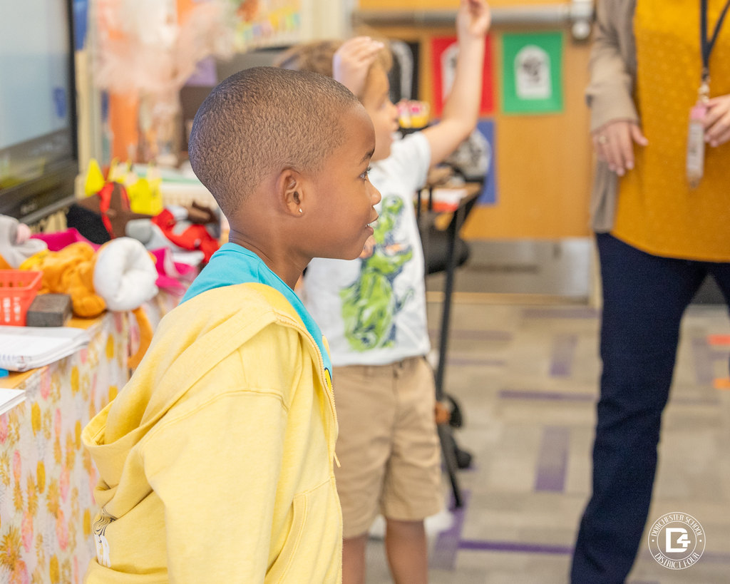 A young boy in a yellow hoodie smiles and raises his hand while standing near the front of the classroom during the counting activity.