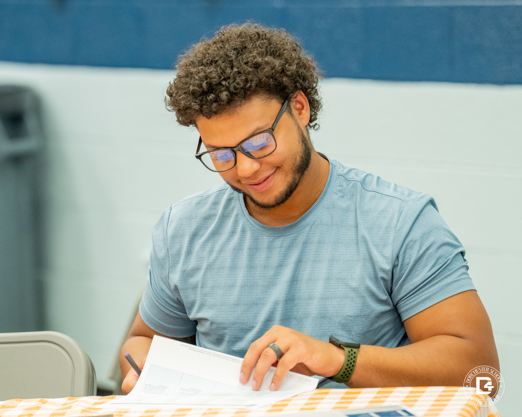 A man with short curly hair and glasses, wearing a light blue shirt, sits at a table with an orange checkered tablecloth. He is smiling and looking down while writing on a piece of paper.