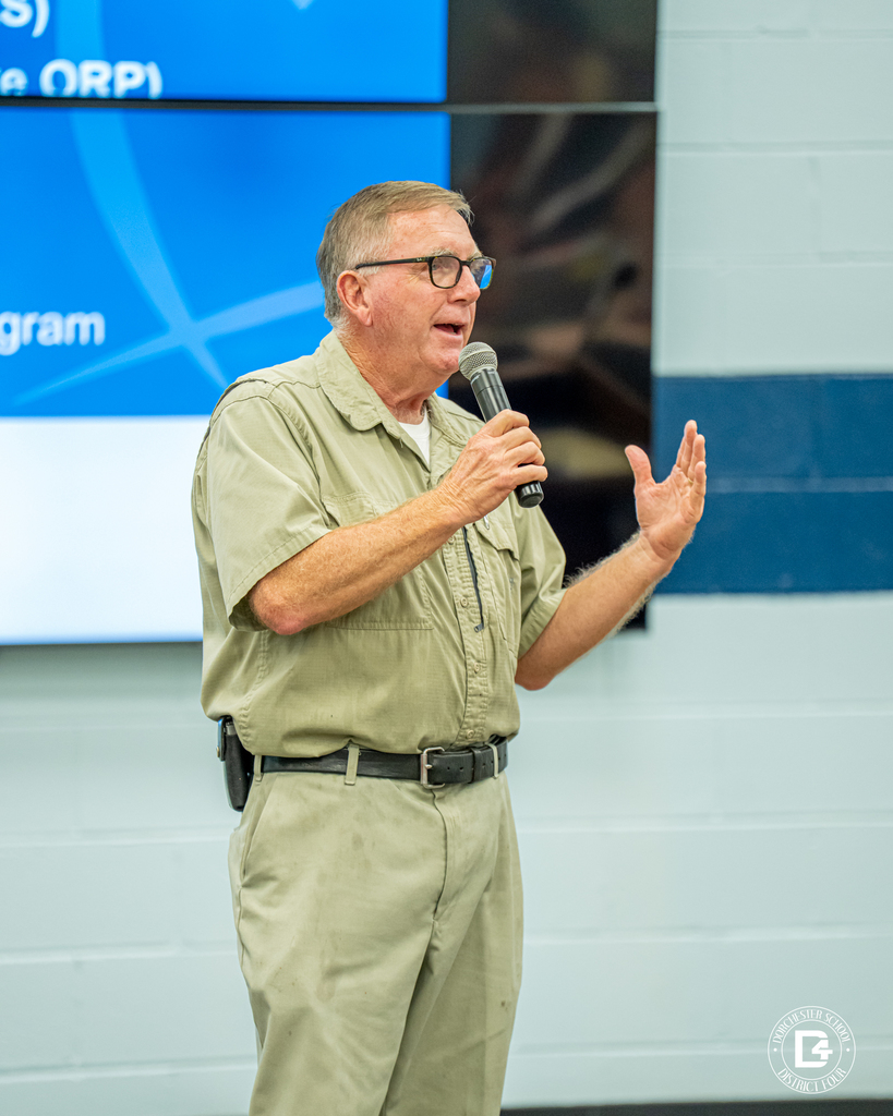 A man in a khaki shirt and glasses holds a microphone while speaking. He gestures with one hand. A large screen with a blue background is visible behind him.