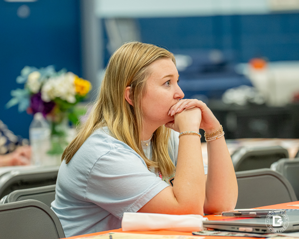 A young woman with long blonde hair, wearing a light blue shirt, sits at a table with her chin resting on her clasped hands. She looks forward attentively, listening. A laptop, papers, and a flower arrangement are visible on the table.