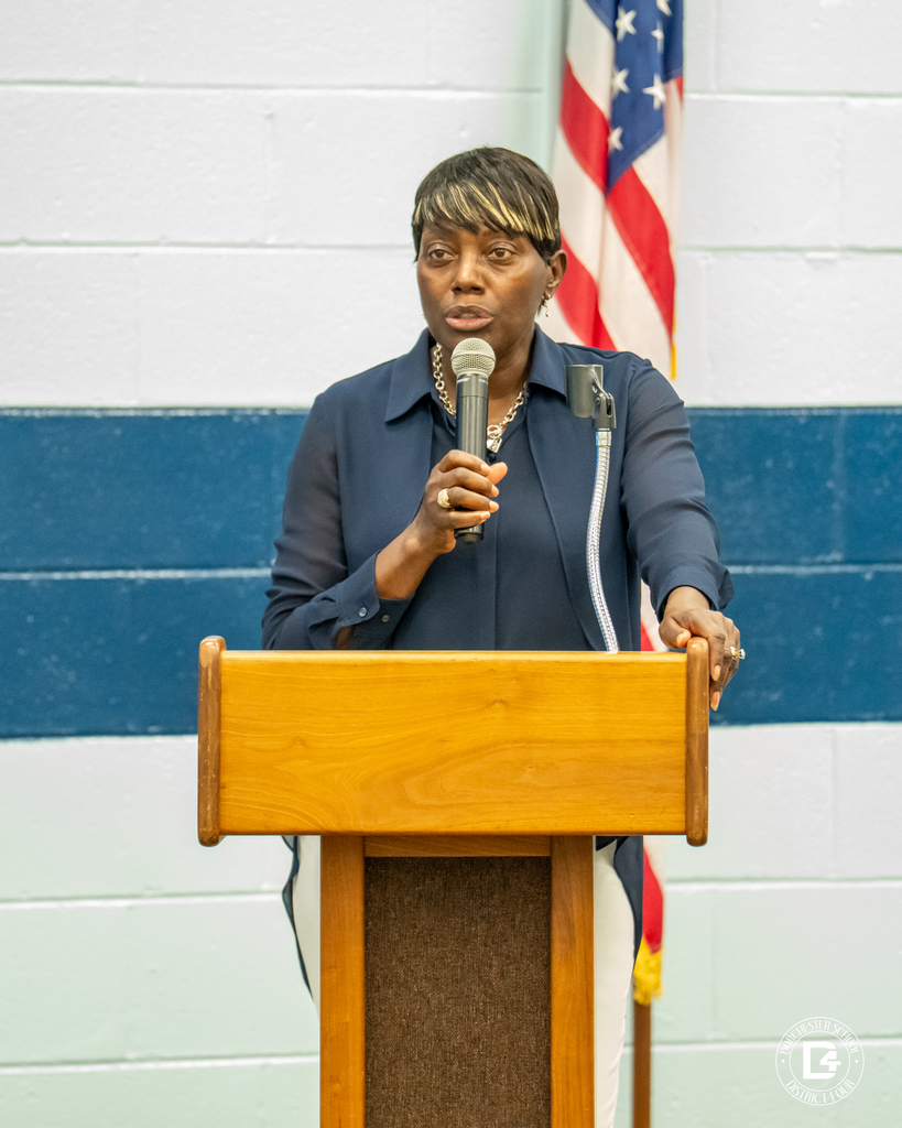 A woman wearing a dark blue blouse stands at a podium, speaking into a microphone. An American flag is visible behind her.