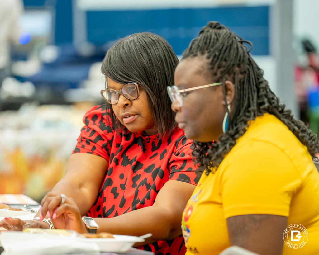 Two women sit side by side at a table. The woman on the left wears glasses and a red patterned blouse, while the woman on the right wears glasses and a yellow shirt. They are focused on something in front of them.