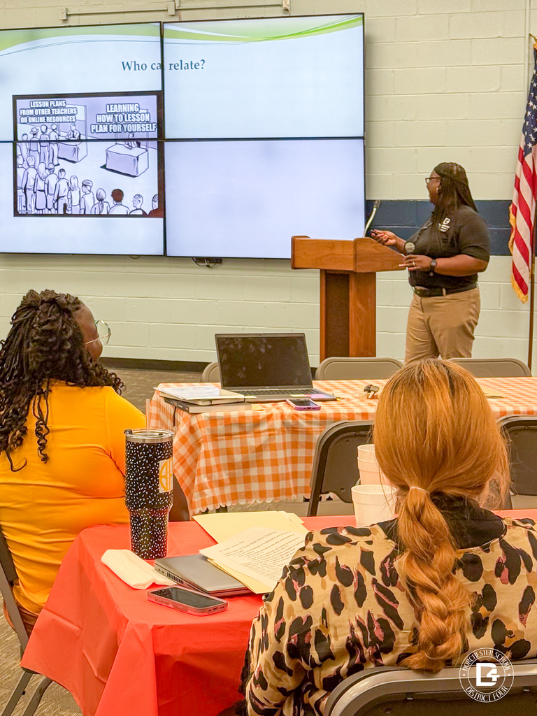 A woman wearing glasses and a black polo shirt with the DD4 logo speaks at a podium in front of a projected presentation slide. Seated attendees listen while facing the screen and podium.