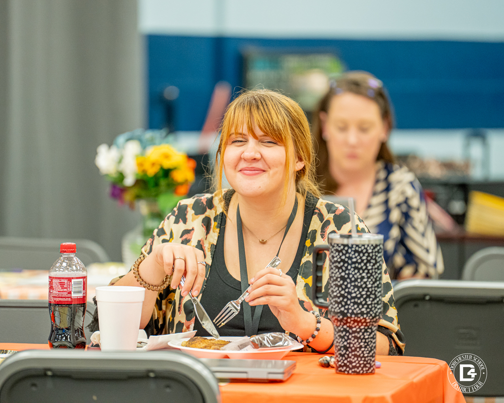 A woman with red hair tied back in a ponytail, wearing a leopard print cardigan, sits at a table with an orange tablecloth. She smiles while holding a fork and eating. Drinks and flowers are visible on the table.