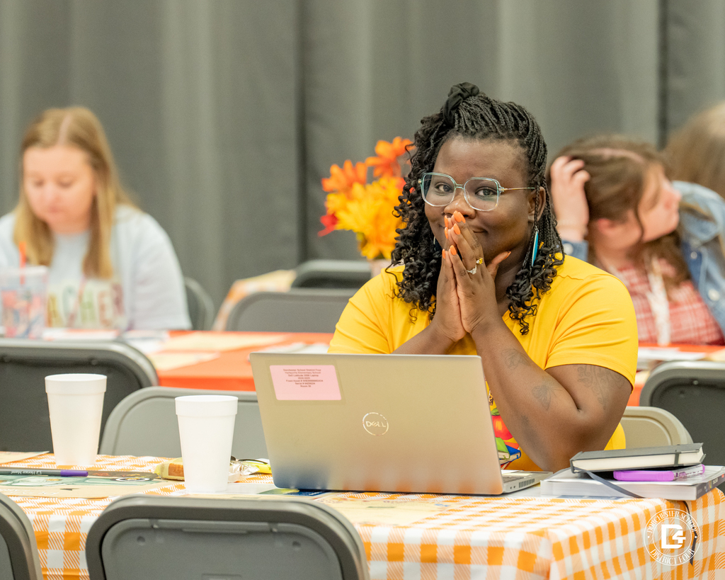 A woman with long braided hair and glasses sits at a table with an orange checkered tablecloth. She is wearing a bright yellow shirt and looking toward the camera with her hands clasped in front of her face, smiling. A laptop and white cups are on the table.