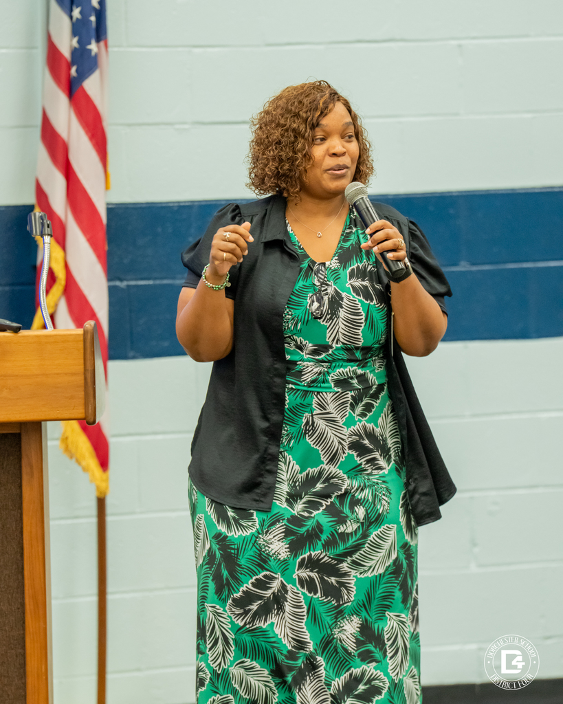 A woman wearing a green and black leaf-patterned dress stands and speaks into a microphone while gesturing with her hand. An American flag is in the background.