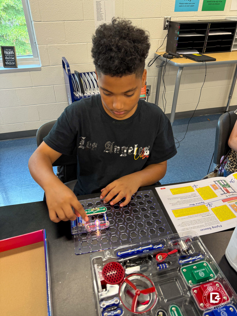 A student in a black t-shirt works on a snap circuits board, pointing to a lit-up bulb on the project while following instructions from the manual.