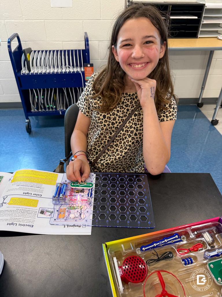 A smiling student in a leopard print shirt poses proudly with her snap circuits project in progress, sitting at a classroom desk with materials spread out.