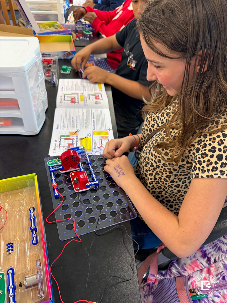 A student in a leopard print shirt follows instructions from a snap circuits manual while assembling a project on their circuit board. Other students in the background are also engaged in building.