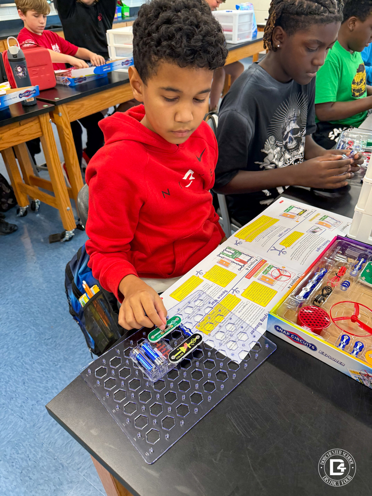 A student in a red hoodie carefully places components onto a snap circuits board while another student beside him works on their project. The snap circuits kit and instruction manual are visible.