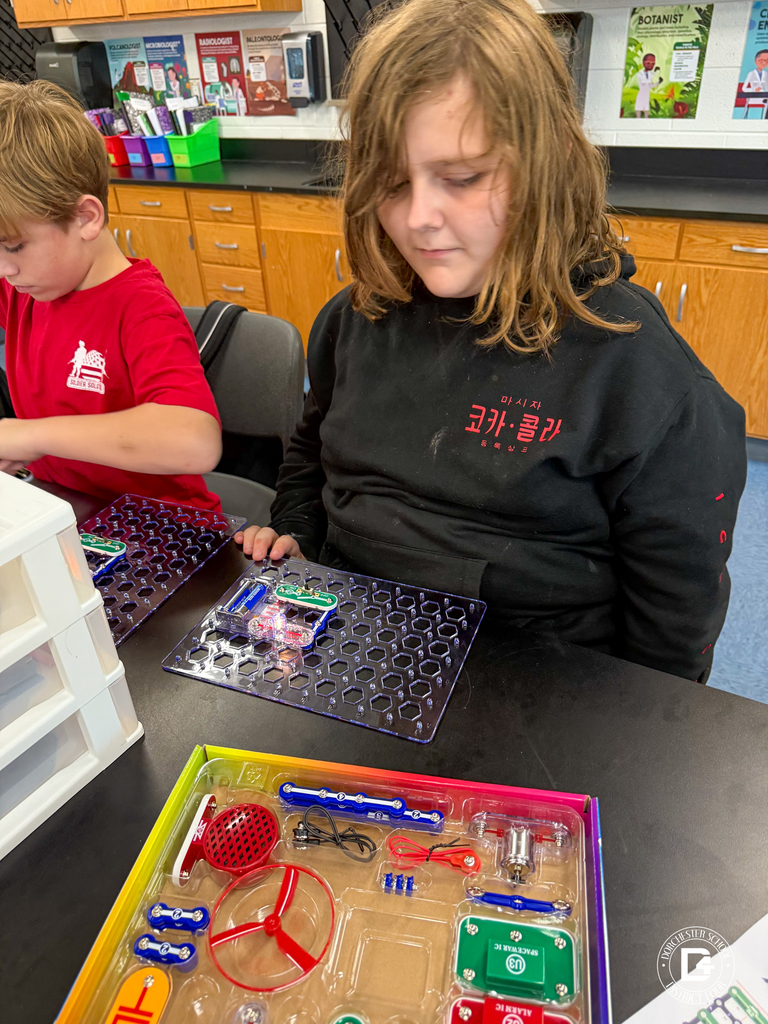 A student in a black hoodie works intently with a snap circuits board, connecting components to create an electronic project. Another student in red is seated beside them working on a similar project.