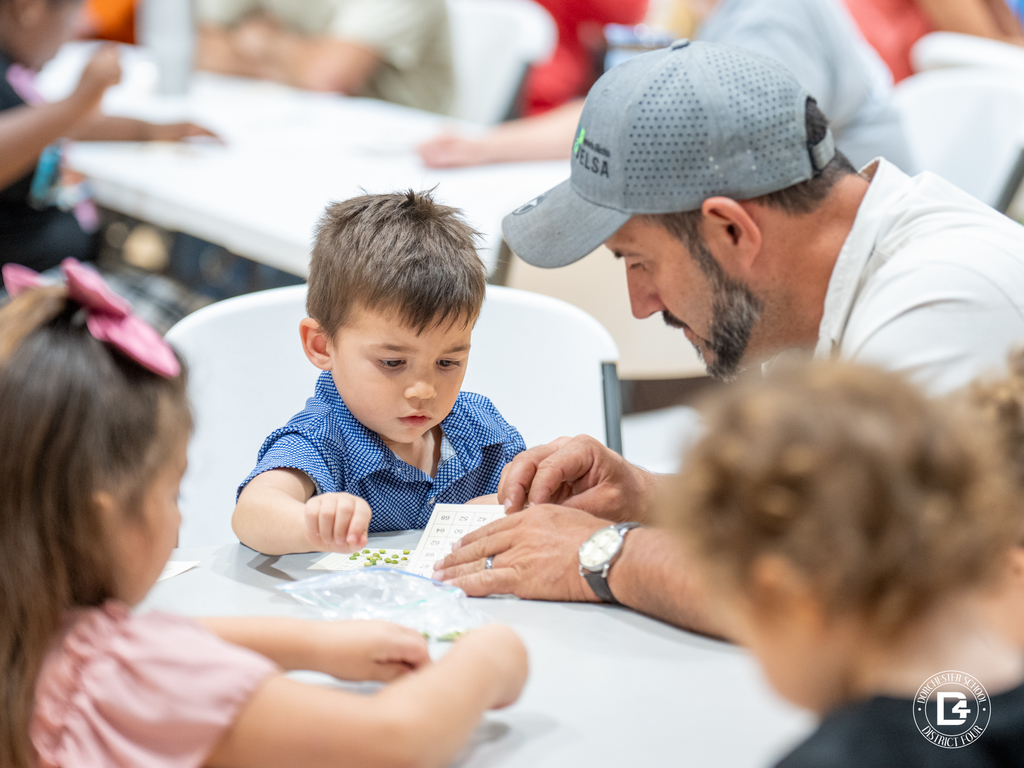 A man helps a young boy check his bingo card, with other children sitting nearby at the same table.