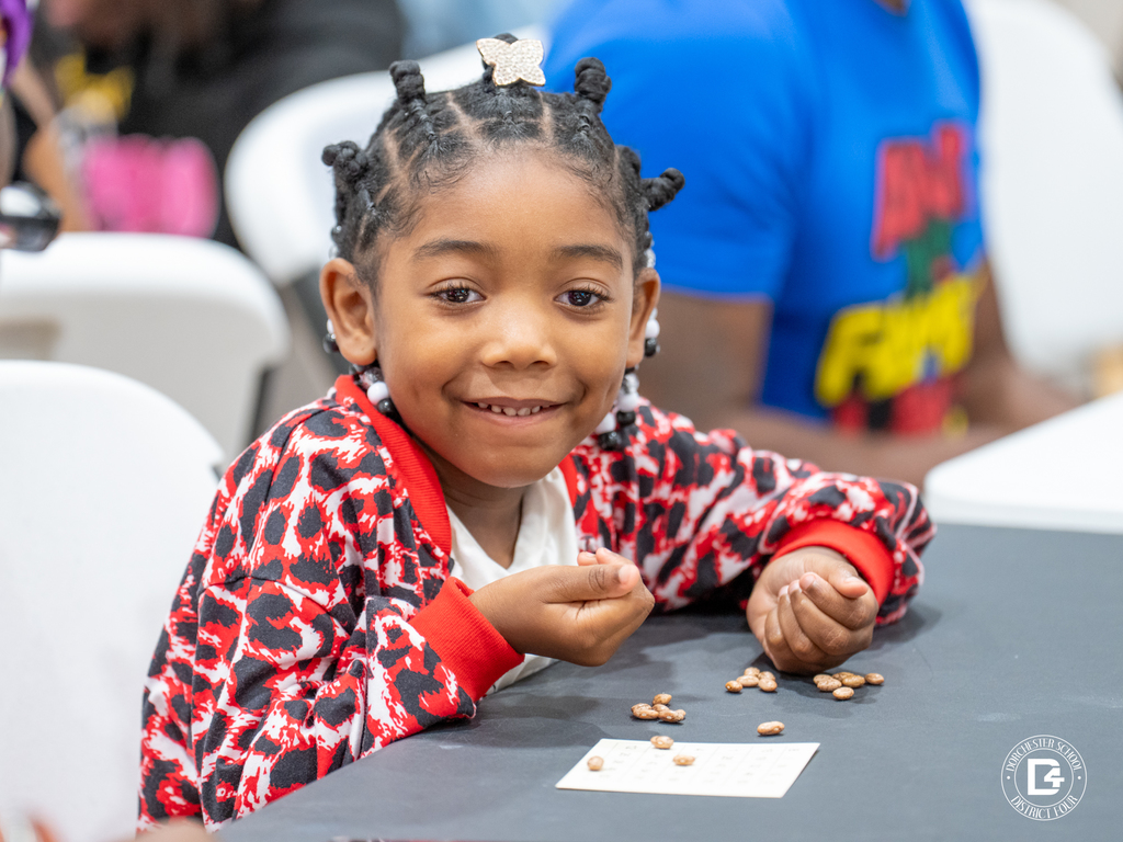 A young girl with braided hair and beads smiles brightly at the camera while holding beans for bingo, seated at a black table.