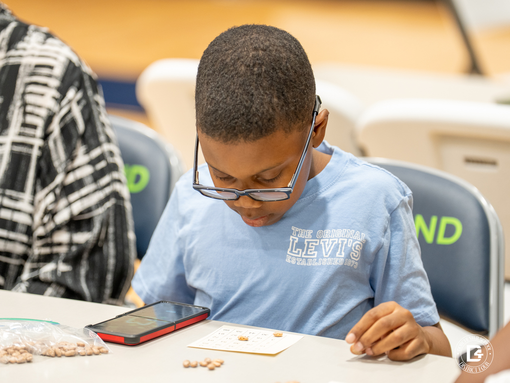 A young boy in glasses studies his bingo card closely while holding a bean marker, with a phone and bag of beans on the table.