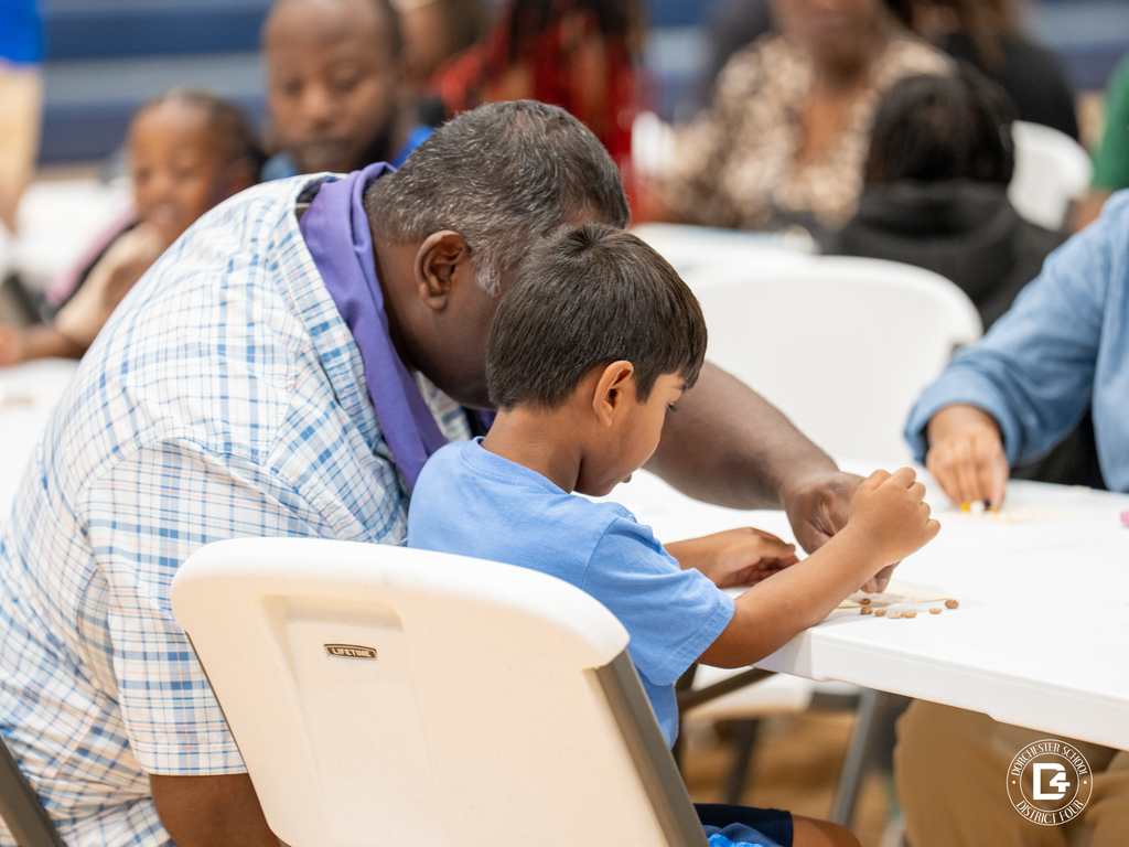 A man leans in close to help a young boy place beans on his bingo card, surrounded by other families playing.
