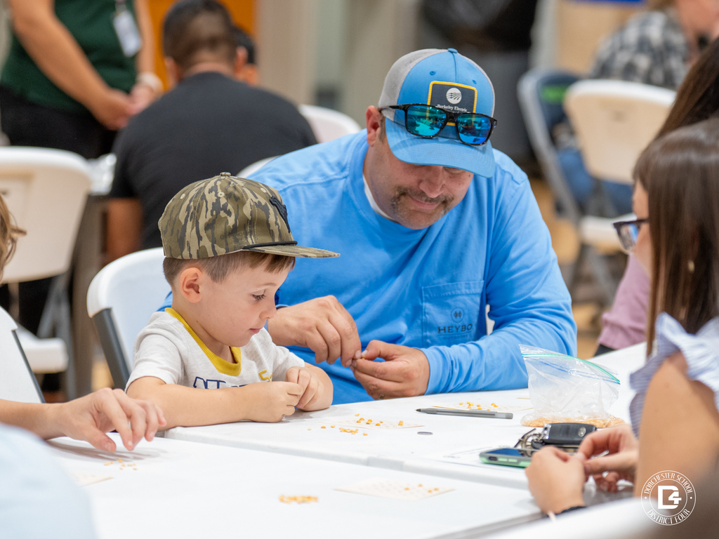 A man in a blue shirt and camo hat helps a little boy mark his bingo card, while other players sit at the same white table.